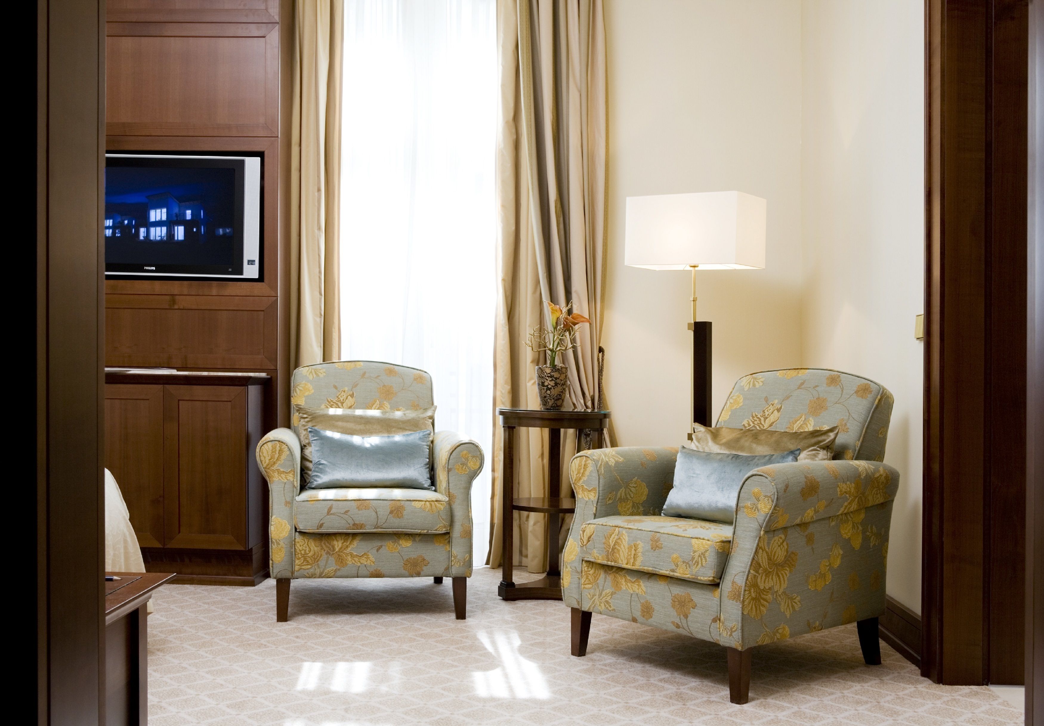A room with two armchairs, a TV mounted on a wooden panel, a lamp, and a vase with flowers.