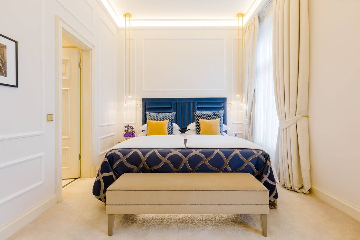 Um quarto com cama, mesas de cabeceira, um banco e cortinas.
