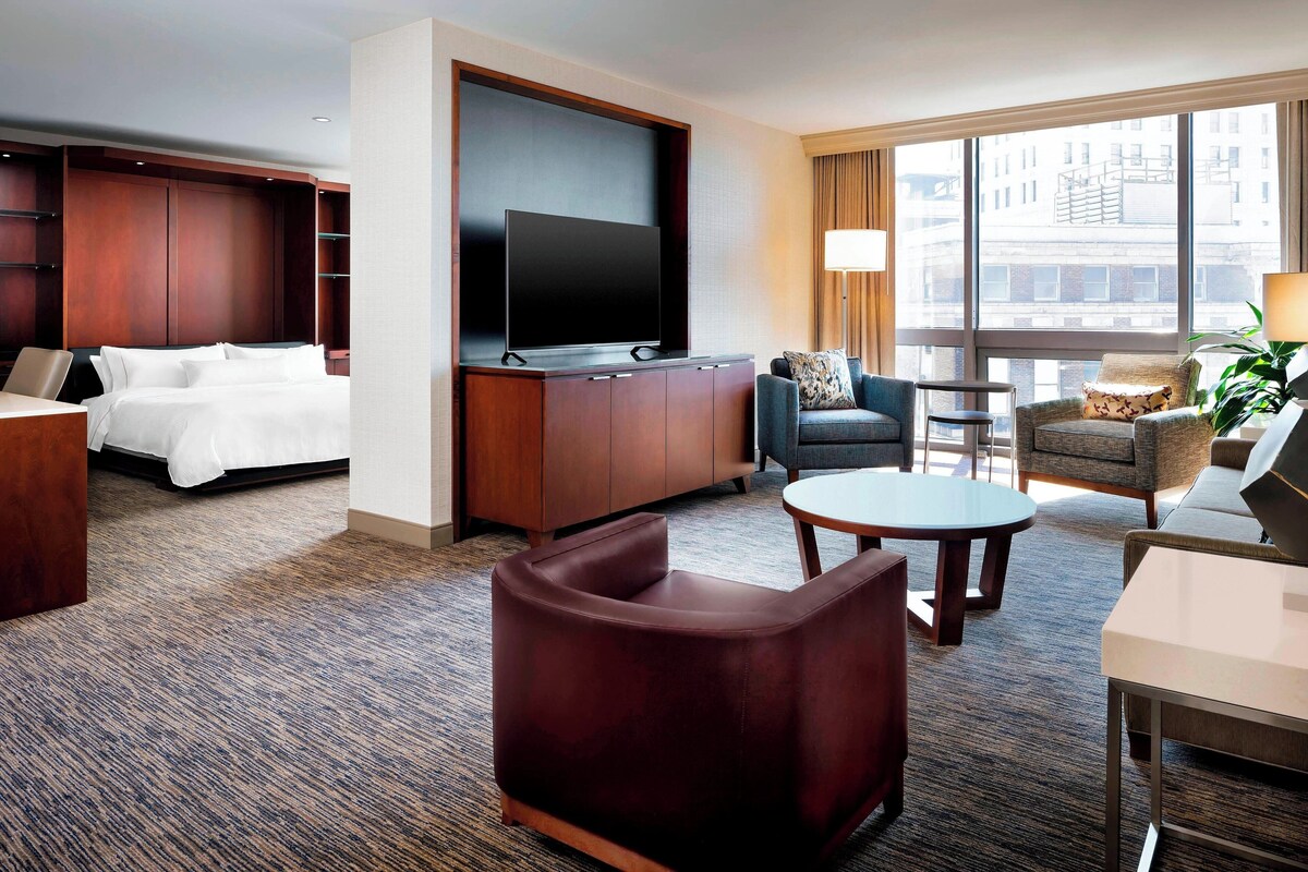 A hotel room with a bed, a TV mounted on a wooden cabinet, a seating area with a sofa and armchairs, and a large window offering a city view.