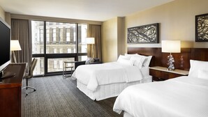 Une chambre d’hôtel avec deux lits, un bureau, une chaise et une grande fenêtre donnant sur des immeubles.