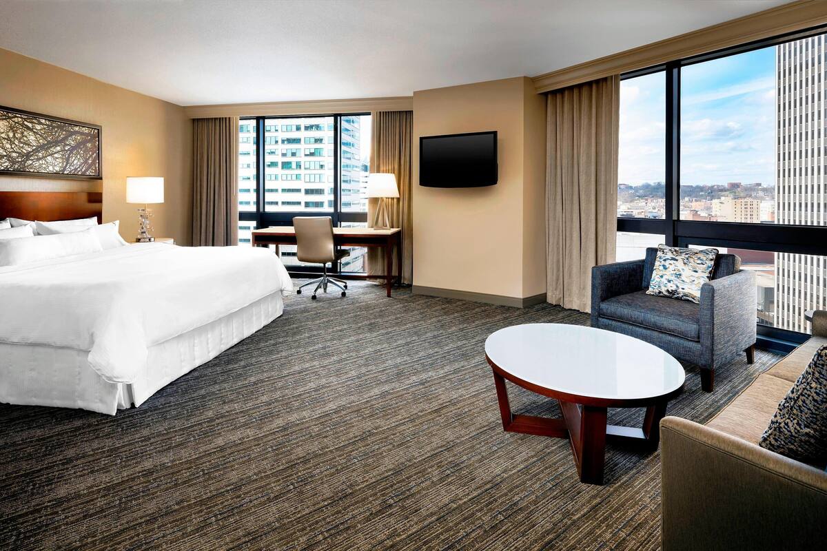 A hotel room with a large bed, a desk, a chair, a sofa, a TV, and a view of the city.