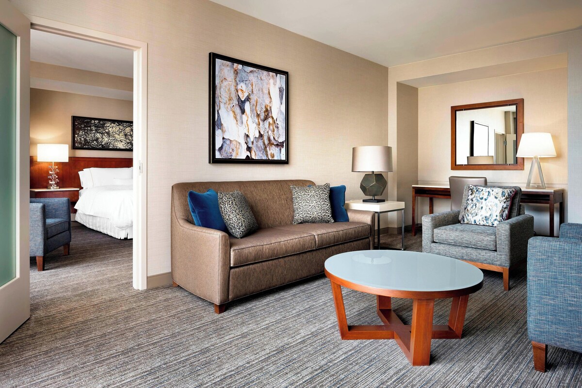 A hotel room with a sofa, armchairs, a coffee table, a bed, and artwork on the walls.