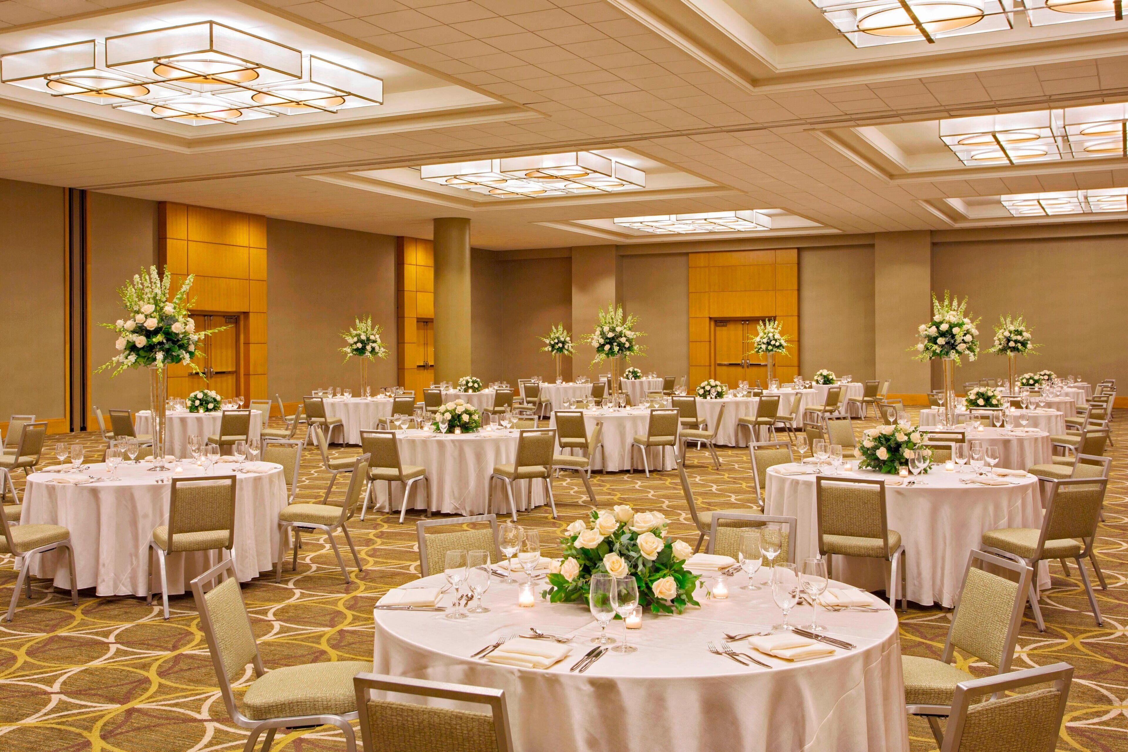 Un salón de banquetes con mesas redondas dispuestas para comer, cada una con manteles blancos y centros de mesa florales.
