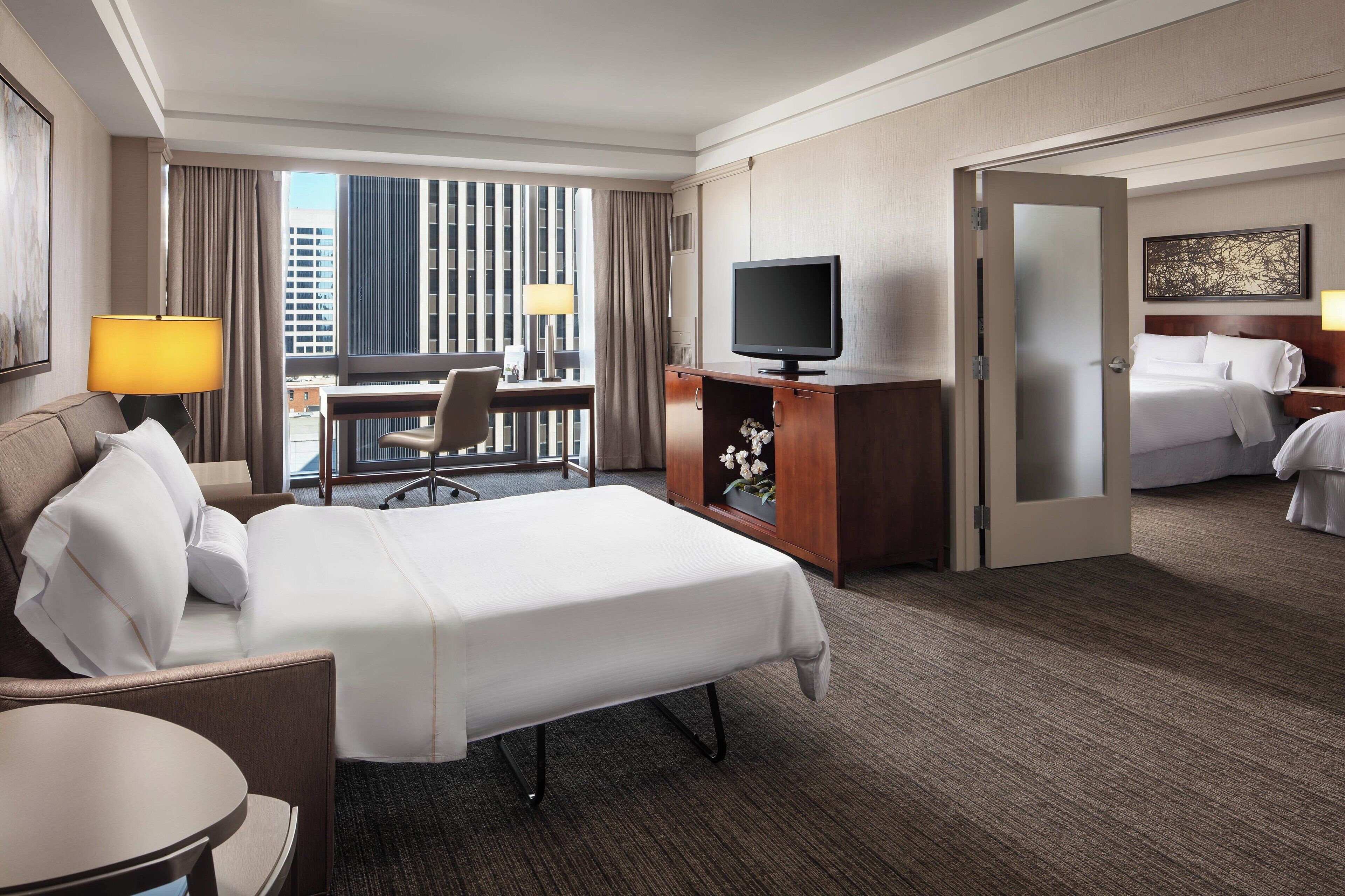 A hotel room with a bed, a desk, a chair, a television, and a view of the city through large windows.