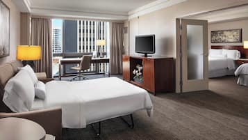 Habitación de hotel con cama, escritorio, silla, televisor y vistas a la ciudad a través de amplias ventanas.