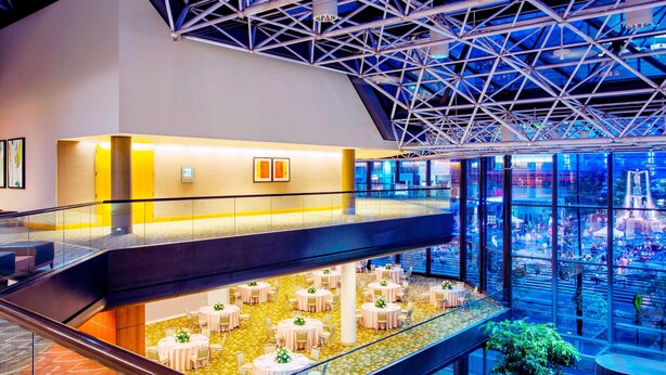 Un atrium à plusieurs niveaux, doté de rampes en verre, d'un plafond haut avec des poutres métalliques, et d'un niveau inférieur avec des tables et des chaises rondes.