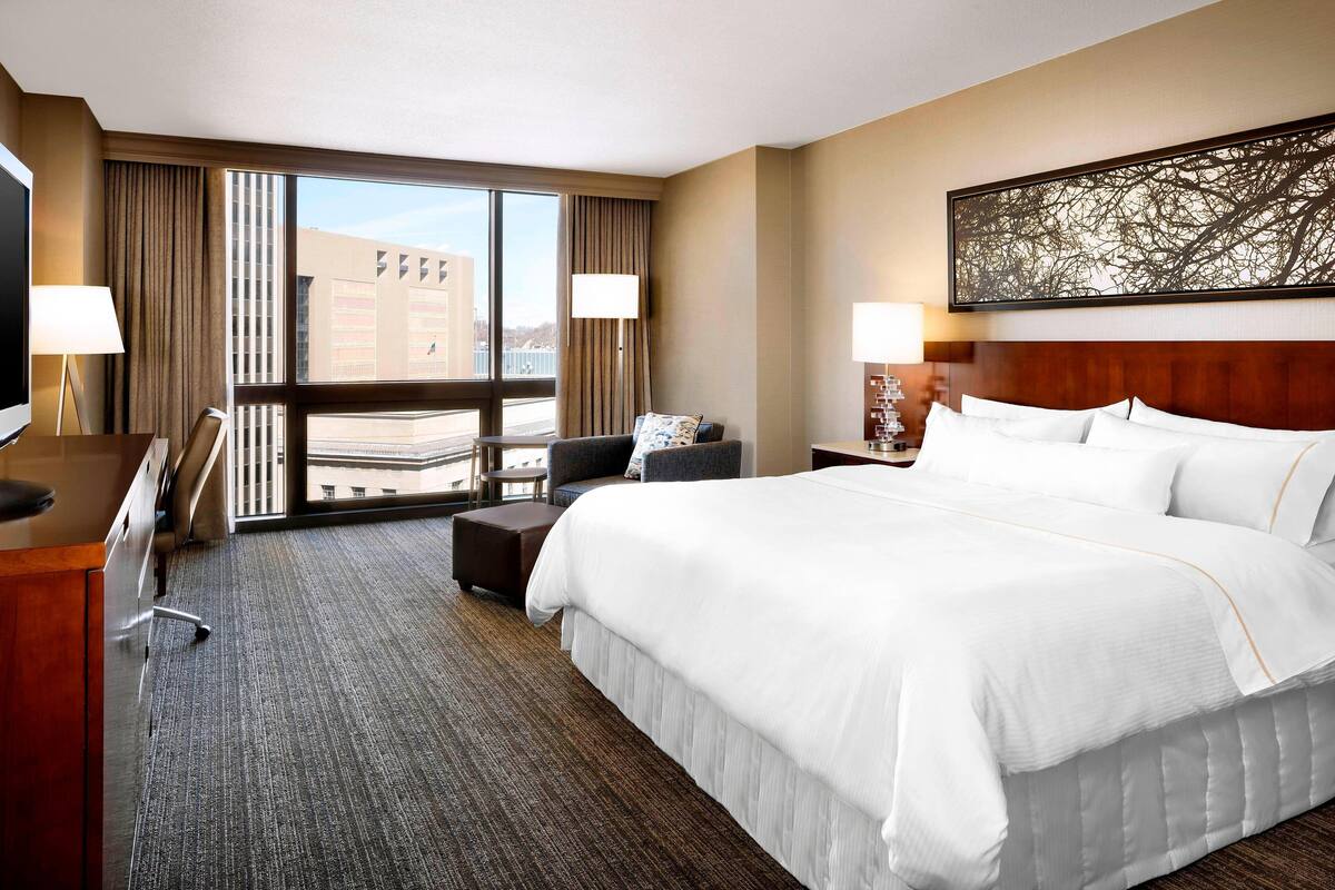 A hotel room with a large bed, a desk, a chair, a small table, and a TV.