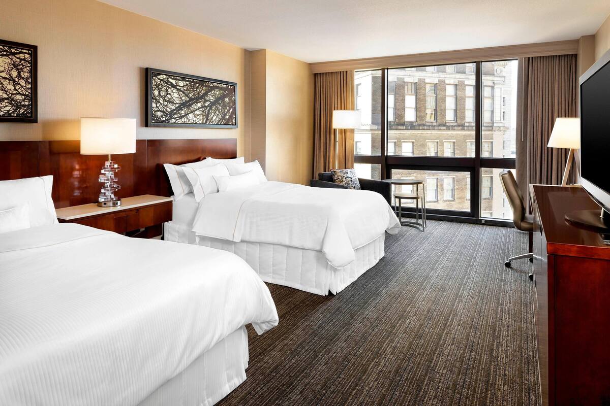 A hotel room with two beds, a desk, a chair, a TV, and a large window with a city view.
