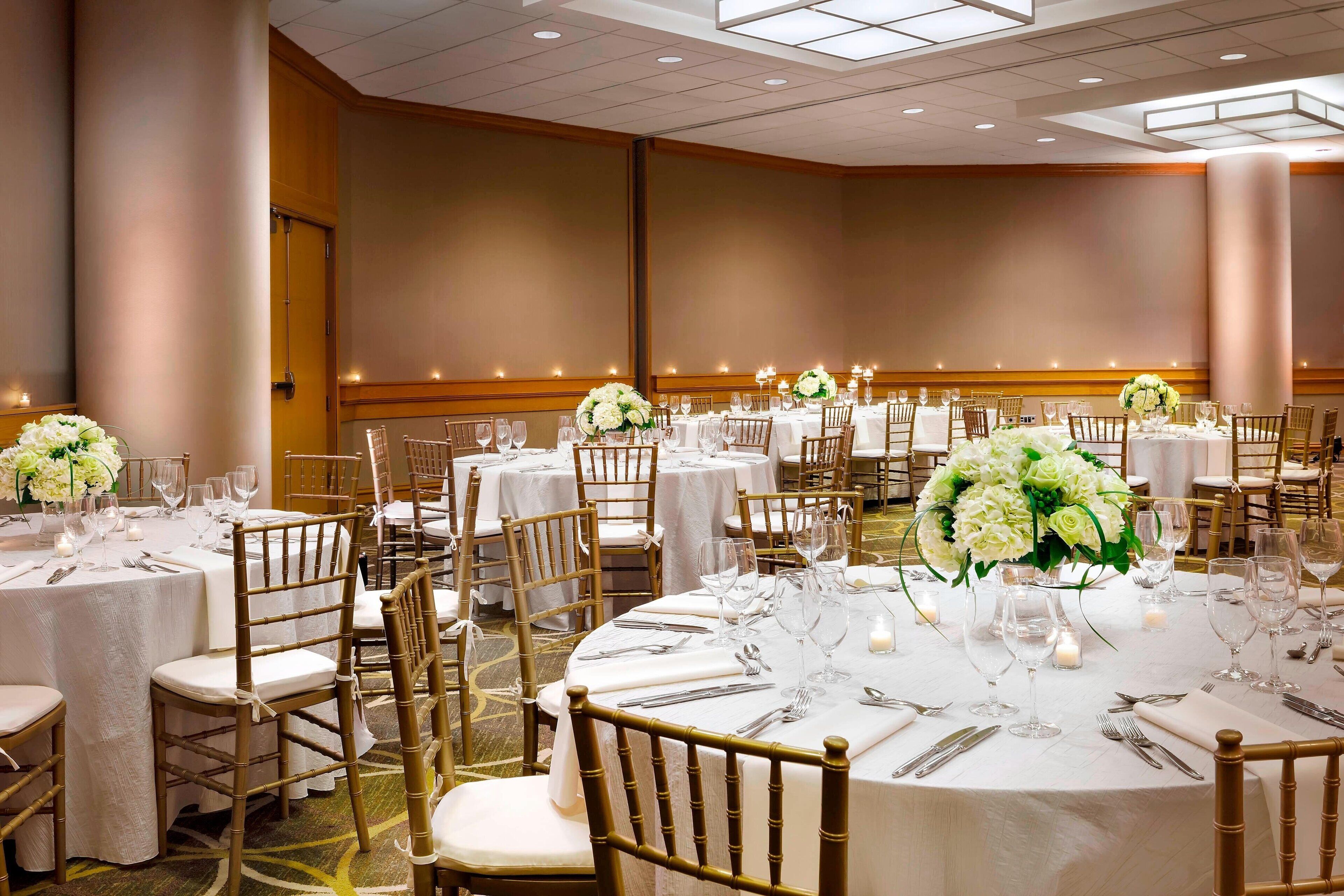 A banquet hall with round tables set for dining, each with white tablecloths, gold chairs, and floral centerpieces.
