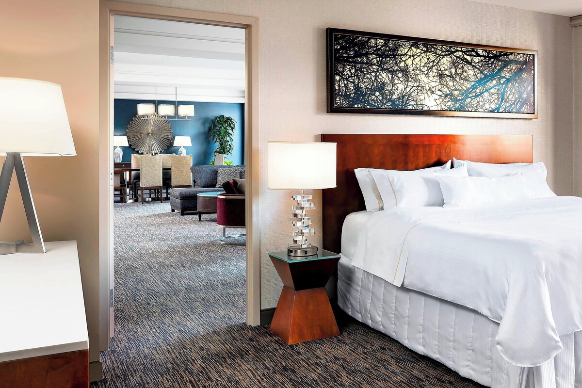 A hotel room with a large bed, a nightstand with a lamp, a modern painting, and a view into the dining area.
