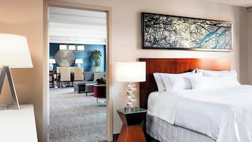 A hotel room with a large bed, a nightstand with a lamp, a modern painting, and a view into the dining area.