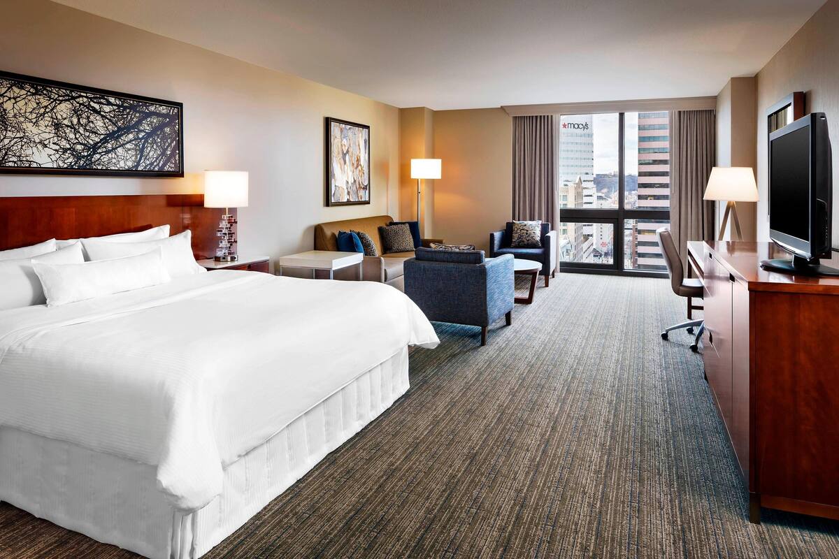 A hotel room with a large bed, a desk with a computer, a chair, and a sofa. There is a window with a view of buildings and a cityscape.