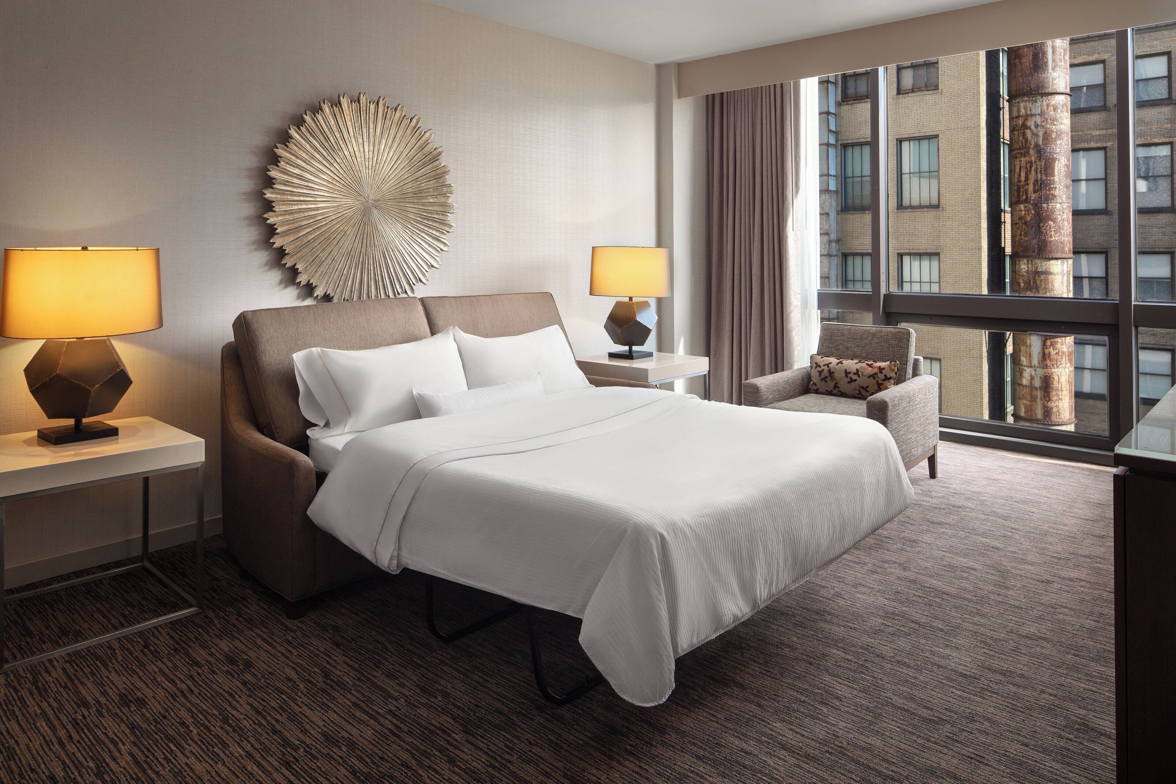 Habitación de hotel con una cama grande, dos mesitas de noche con lámparas, un adorno en la pared, vista de un edificio a través de una ventana y una silla.