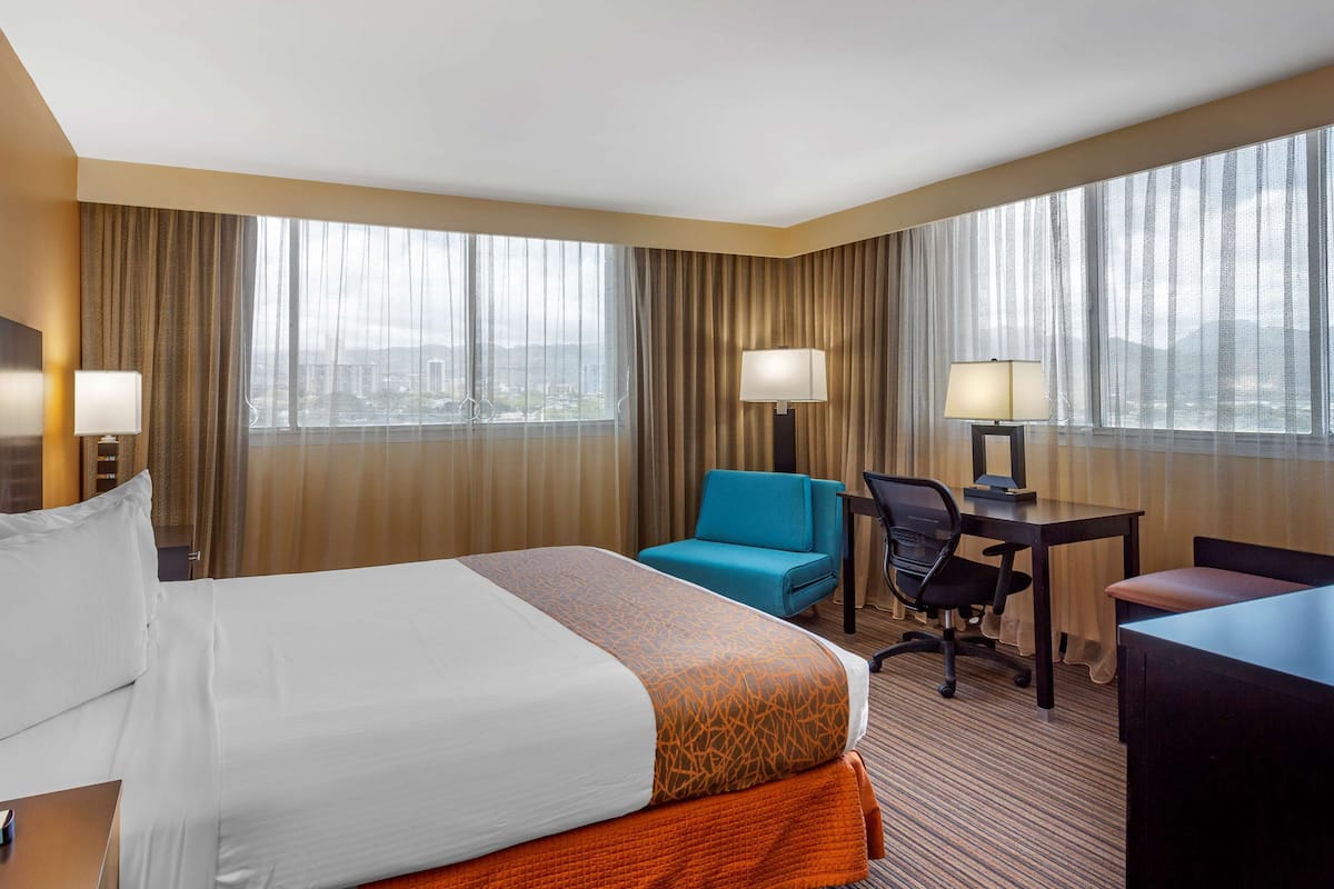Une chambre d’hôtel avec un grand lit, un bureau, une chaise et une vue sur la ville grâce à de grandes fenêtres.