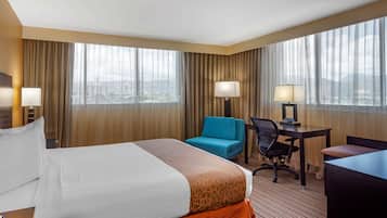 A hotel room with a large bed, a desk, a chair, and a view of the city through large windows.