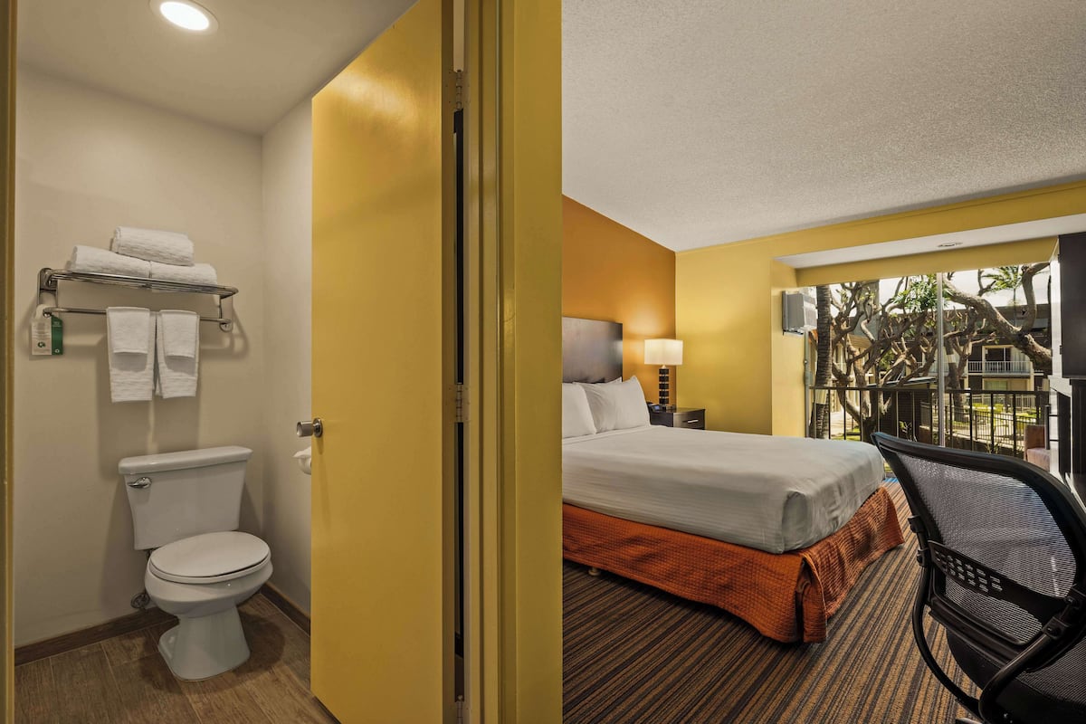 Une chambre d’hôtel avec des toilettes, un lit recouvert d’une couverture orange, une chaise et une table de chevet avec une lampe.
