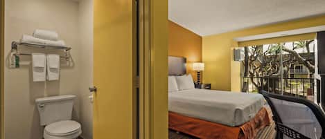 A hotel room with a toilet, a bed with an orange bedspread, a chair, and a nightstand with a lamp.