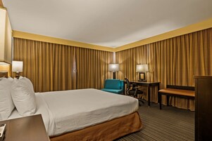 A hotel room with a large bed, a desk, a chair, and a desk lamp.