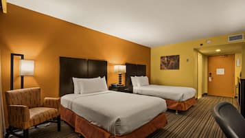 A hotel room with two beds, a chair, a desk, and a lamp.