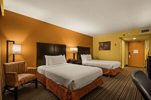 Quarto de hotel com duas camas, uma cadeira, uma escrivaninha e um abajur.