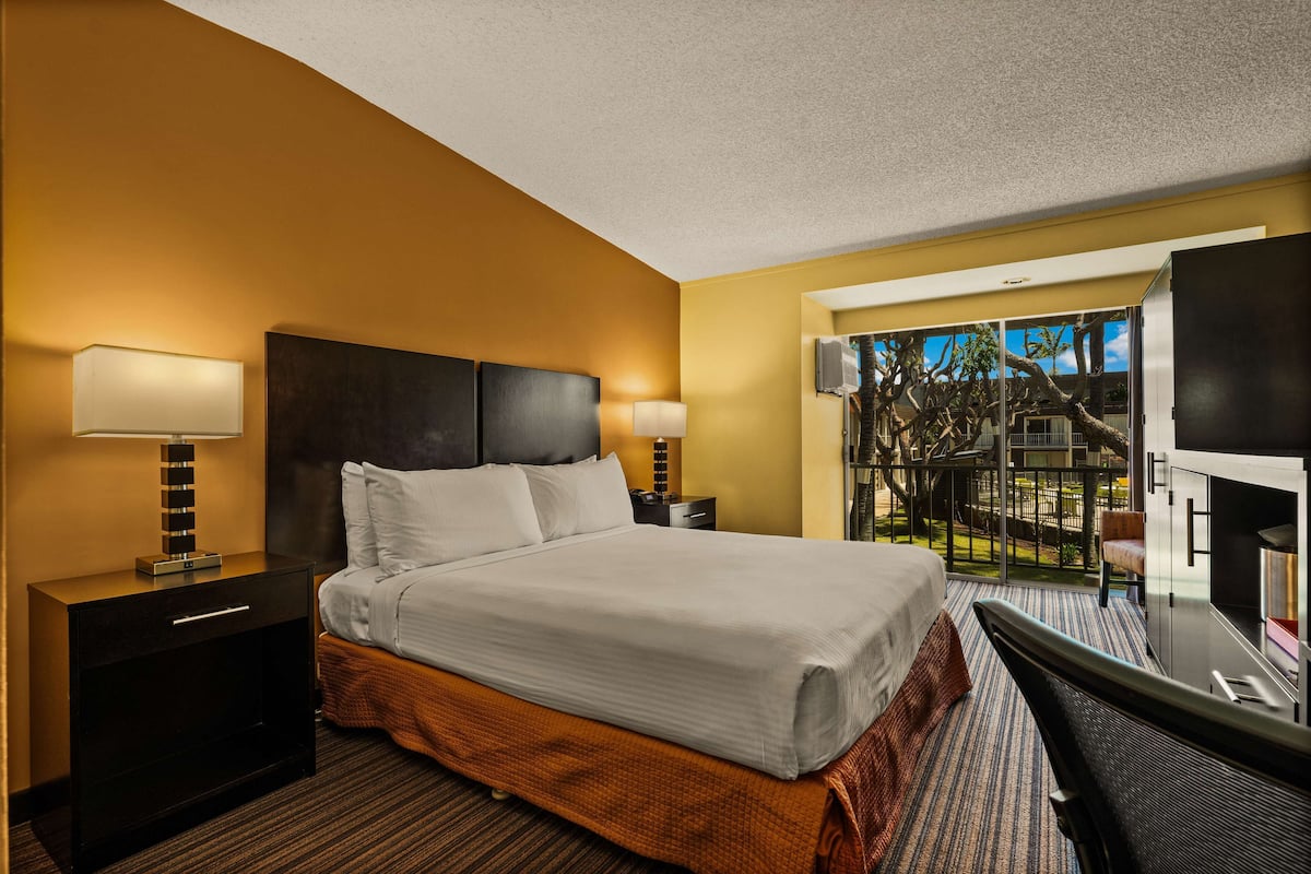 Une chambre d’hôtel avec un grand lit, deux tables de chevet avec des lampes, une télévision et un balcon offrant une vue sur les arbres et le ciel.