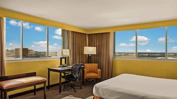 Ein Hotelzimmer mit Schreibtisch, Stuhl und Bett, große Fenster mit Blick auf die Stadt und eine Lampe auf dem Schreibtisch.