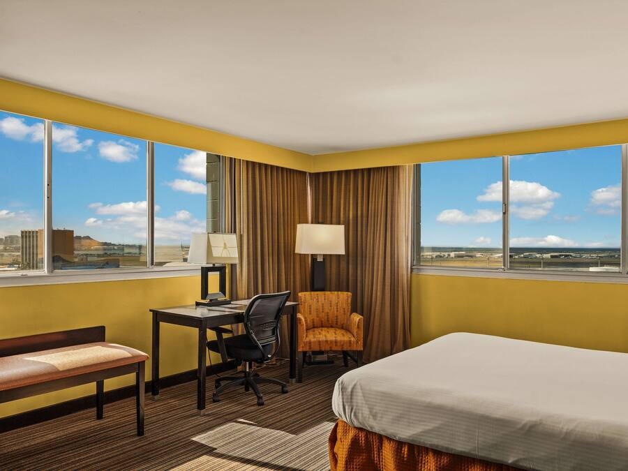 Ein Hotelzimmer mit Schreibtisch, Stuhl und Bett, große Fenster mit Blick auf die Stadt und eine Lampe auf dem Schreibtisch.