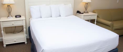 A neatly made bed with white linens and pillows, flanked by two bedside tables with lamps and a phone. A beige sofa is positioned to the right.