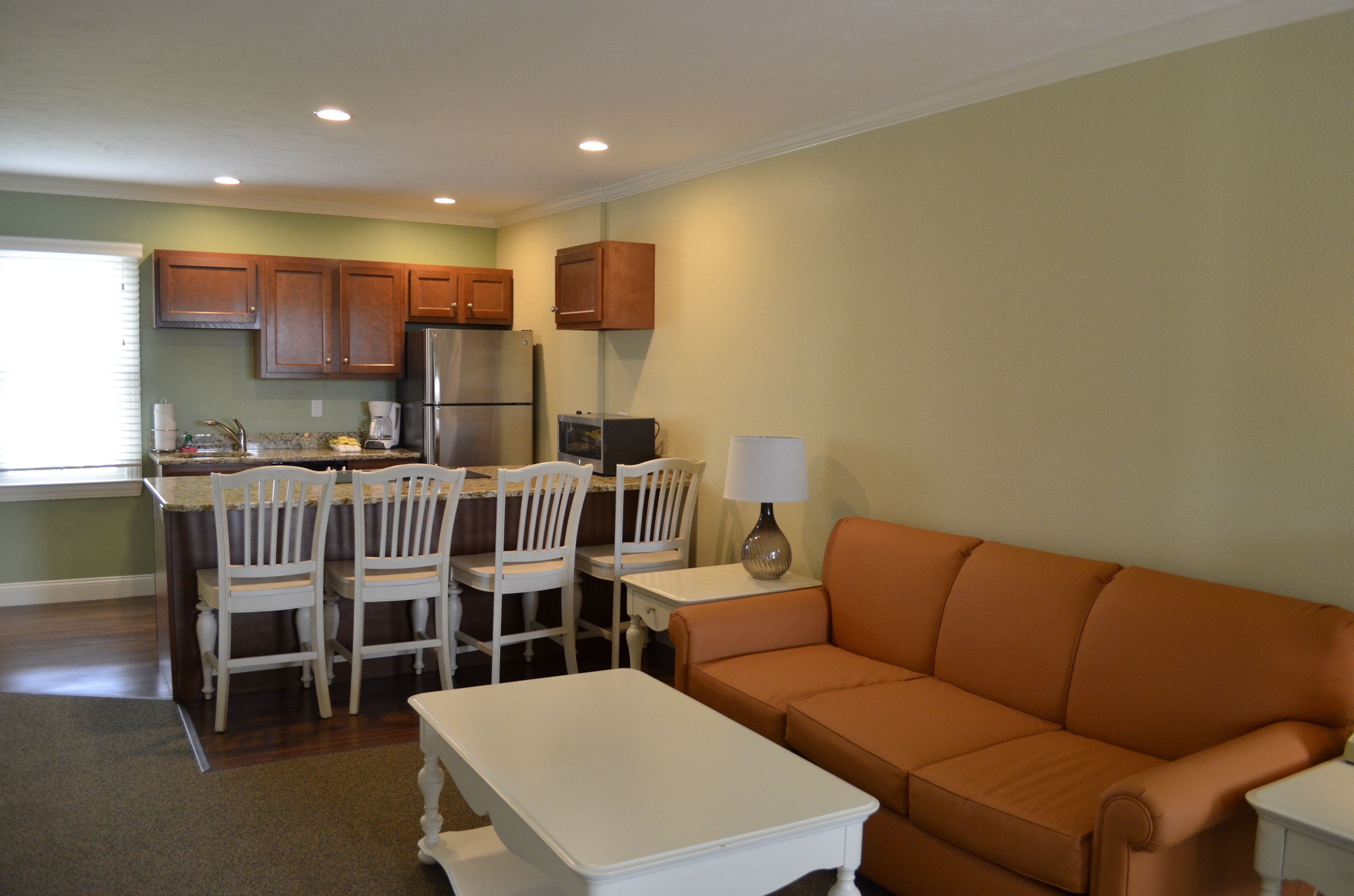 A compact living space with a kitchen area, dining table, chairs, a sofa, and a small table with a lamp.
