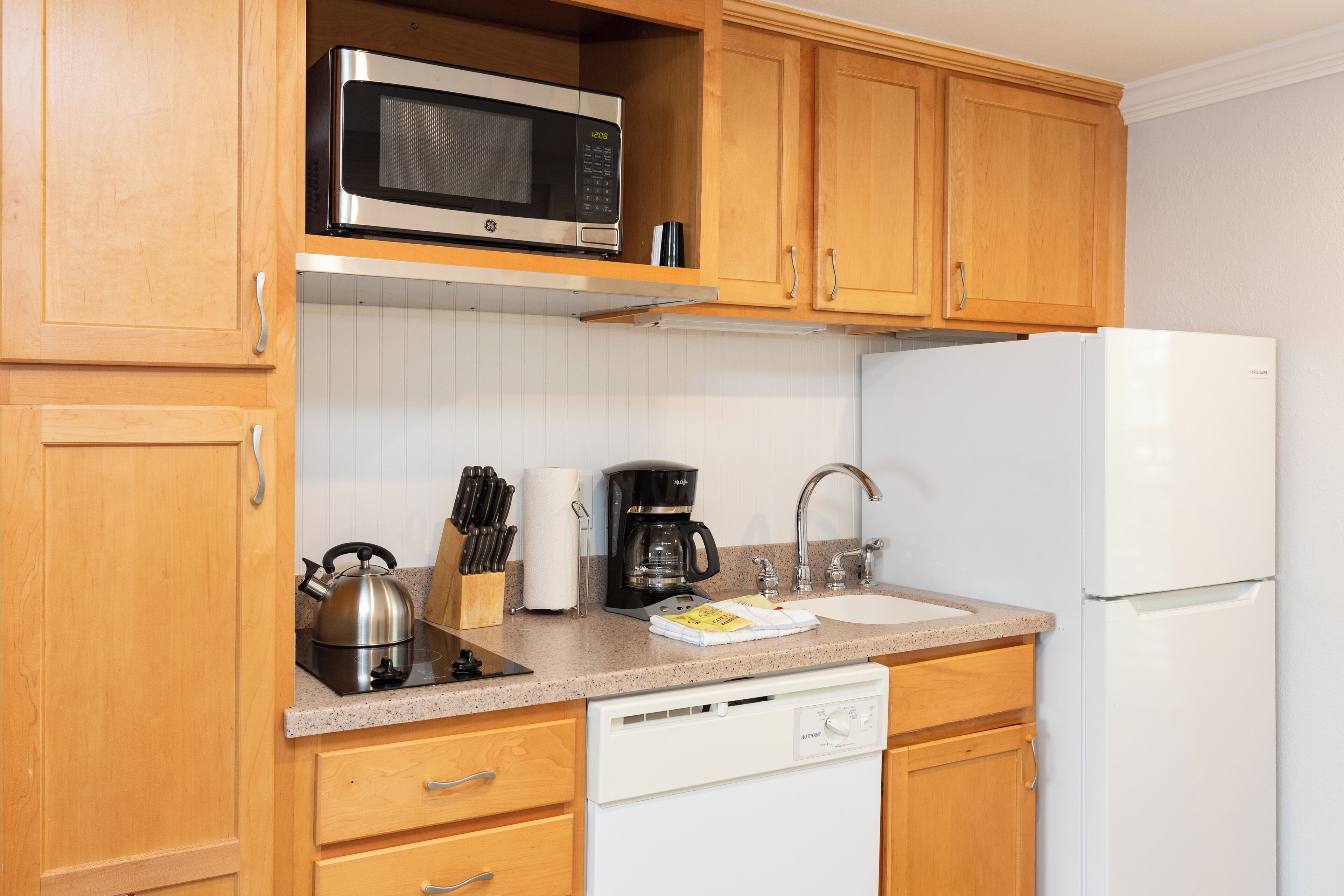 A kitchen with wooden cabinets, a stainless steel microwave, a coffee maker, a kettle, and a white dishwasher.