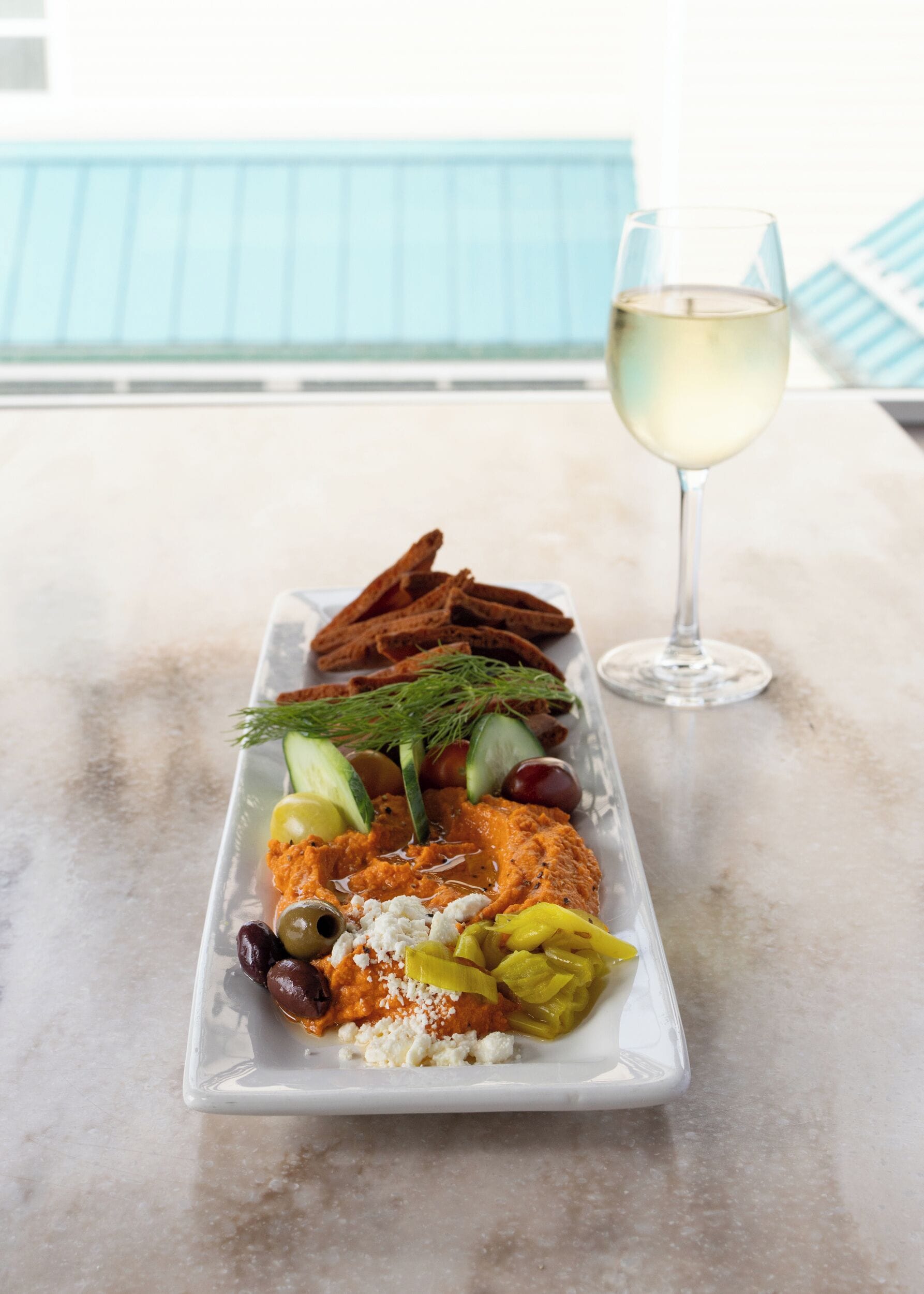 A rectangular white plate with various food items and a glass of white wine on a marble table.