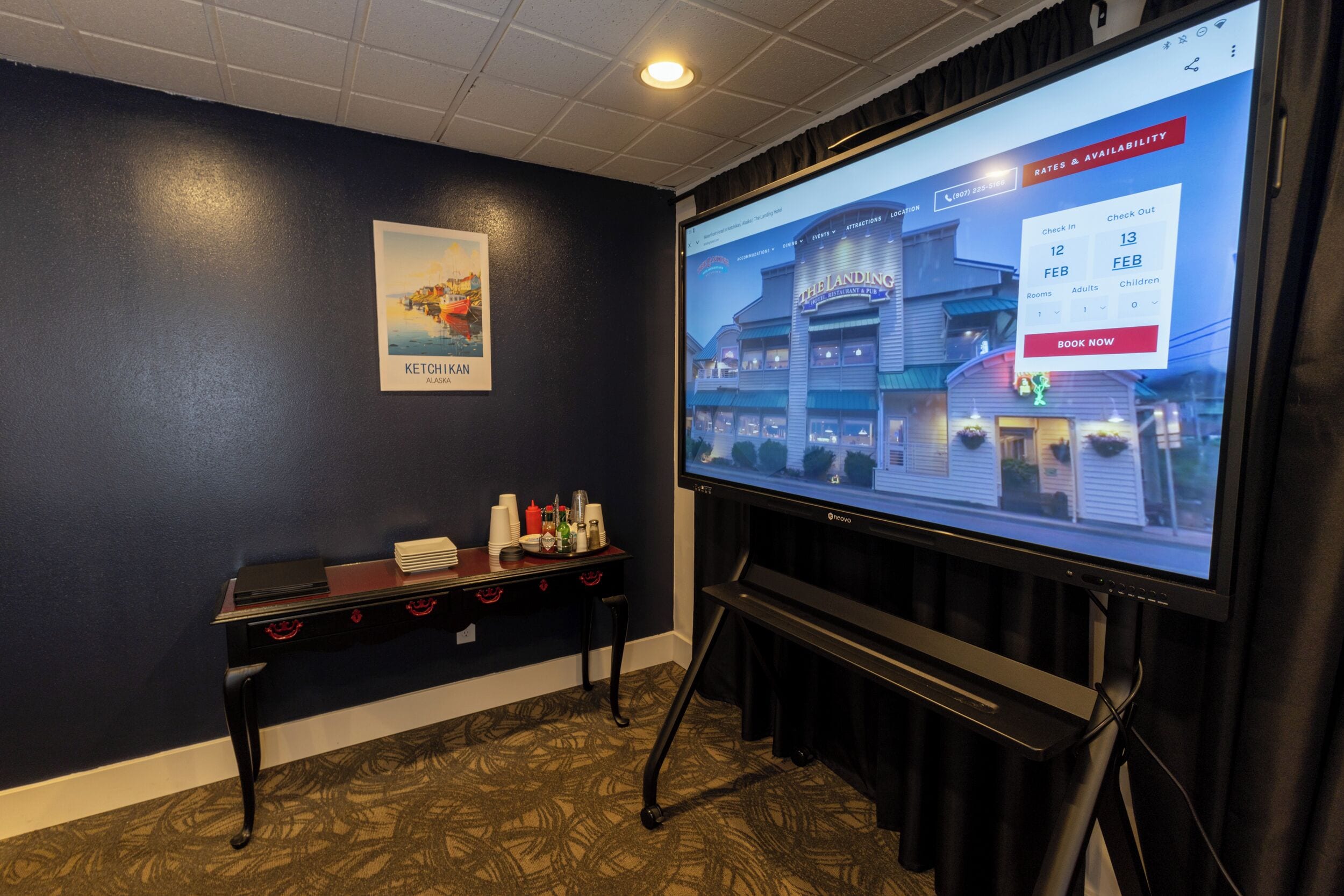 A hotel room with a large screen displaying a booking interface, a small table with a stack of black folders, and a framed picture on the wall.