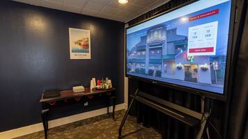 A hotel room with a large screen displaying a booking interface, a small table with a stack of black folders, and a framed picture on the wall.