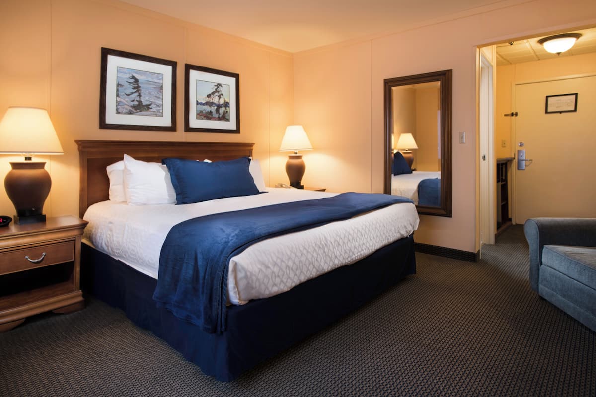 A hotel room with a large bed, two bedside lamps, a nightstand, a dresser, a mirror, and a door leading to another room.