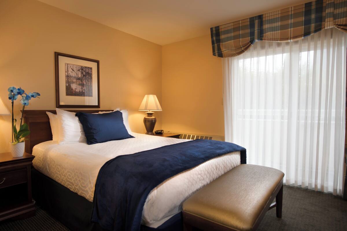 A hotel room with a large bed, a bedside table with a lamp, a framed picture on the wall, and a window with sheer curtains.