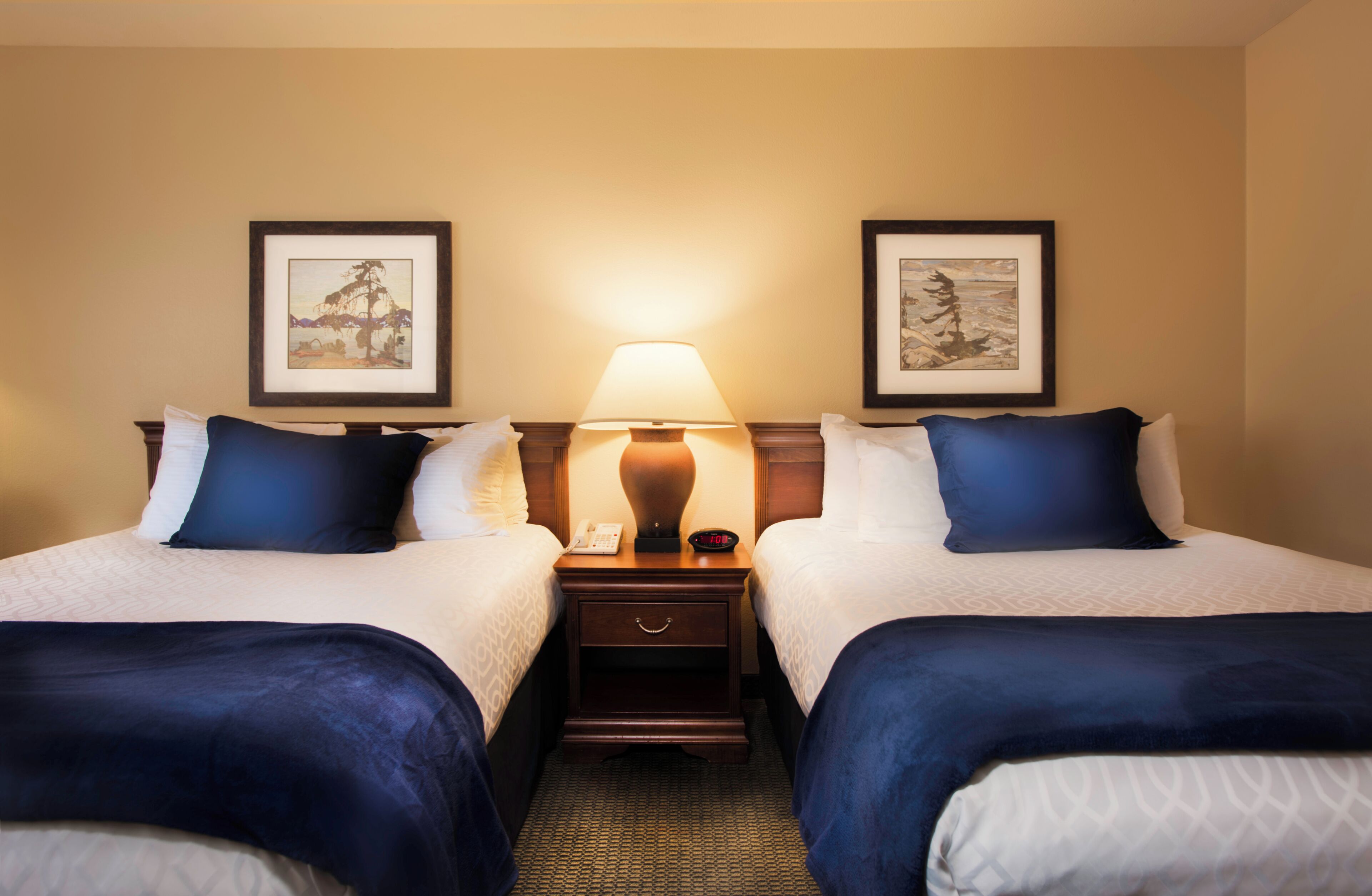 A hotel room with two beds, a nightstand with a lamp, and framed artwork on the walls.