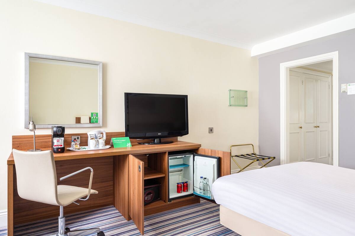 A hotel room with a bed, a desk with a television, a mini-fridge, and a chair.