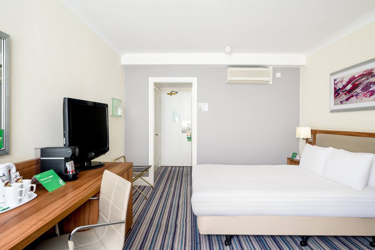 A hotel room with a bed, a desk with a computer, a chair, a television, and a door leading to another room.