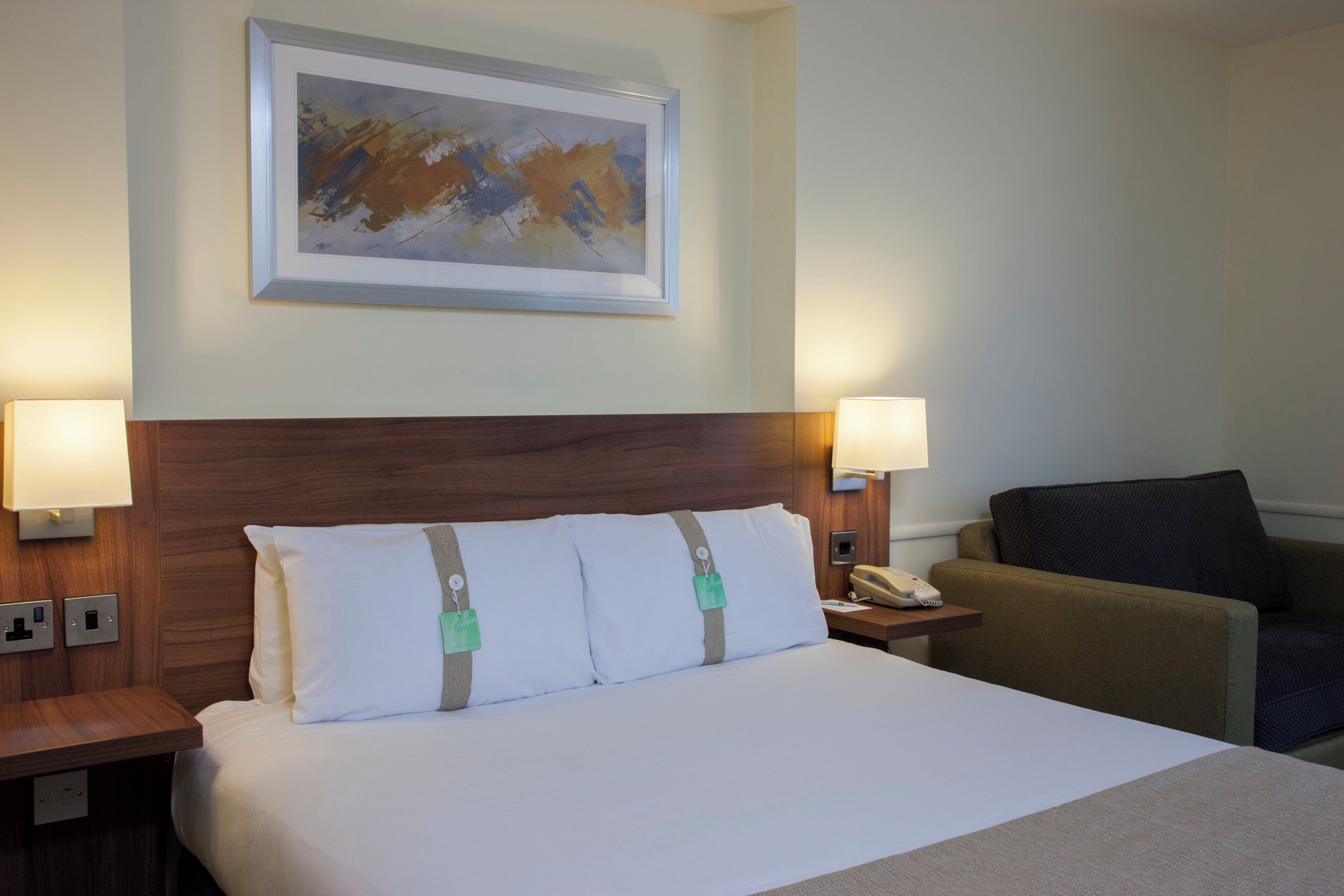 A hotel room with a large bed, two bedside lamps, a telephone, a chair, and a painting on the wall.
