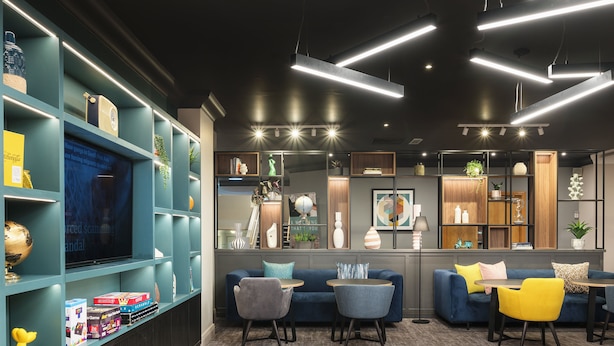 A modern living room with a dark blue sofa, two armchairs, and a small table. There are shelves with decorative items and a flat-screen TV. - Holiday Inn Leeds Wakefield M1 Jct.40 by IHG (Ossett)