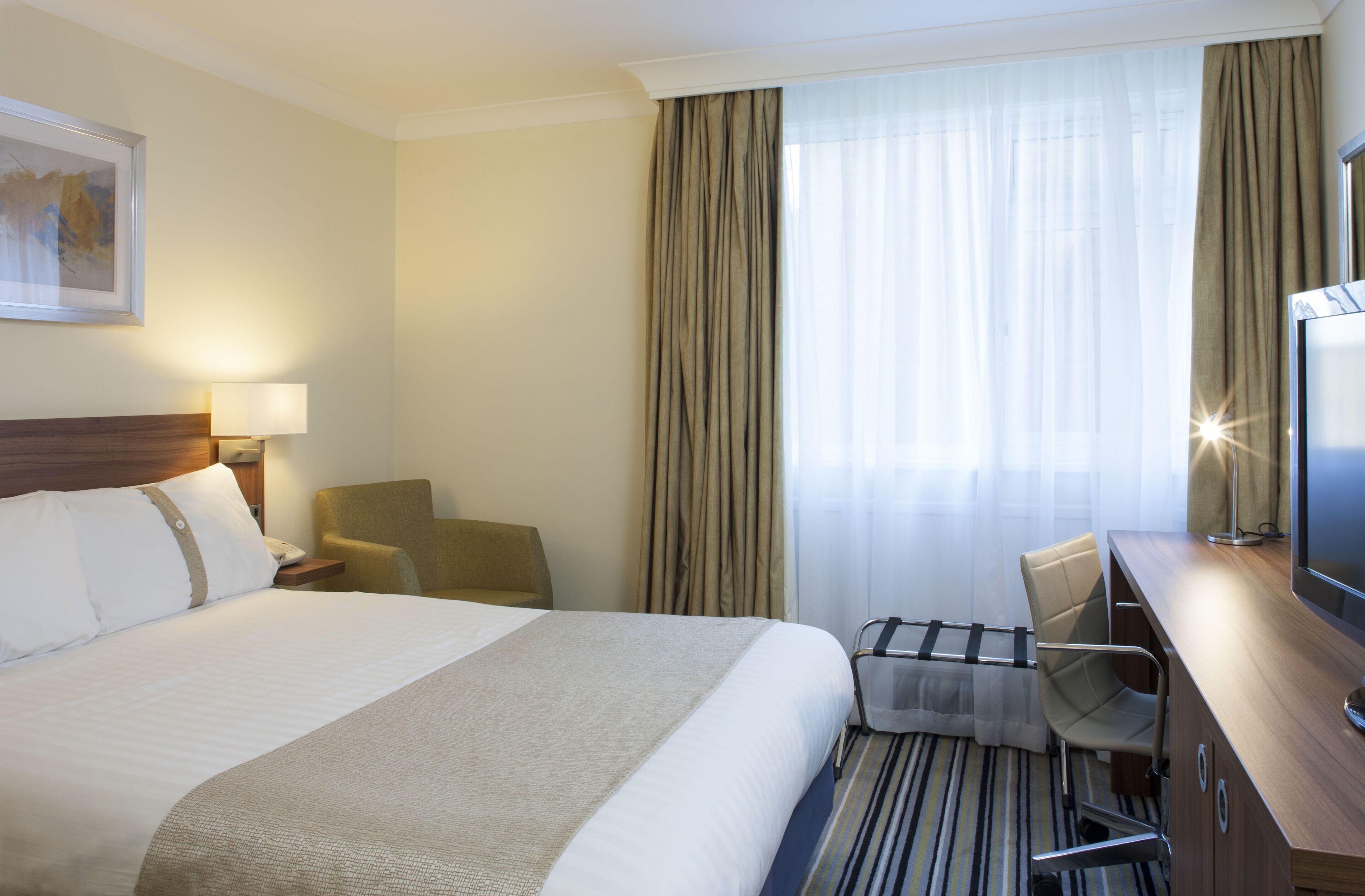 A hotel room with a large bed, a desk with a chair, a television, and a window with curtains.