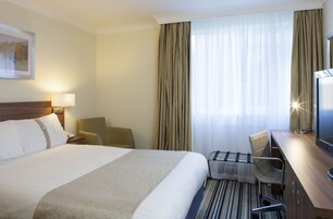 Quarto de hotel com uma cama grande, uma escrivaninha com cadeira, televisão e janela com cortinas.