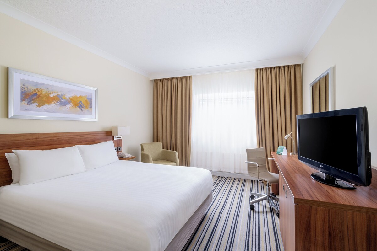 A hotel room with a large bed, a desk with a flat-screen TV, a chair, and a painting on the wall.