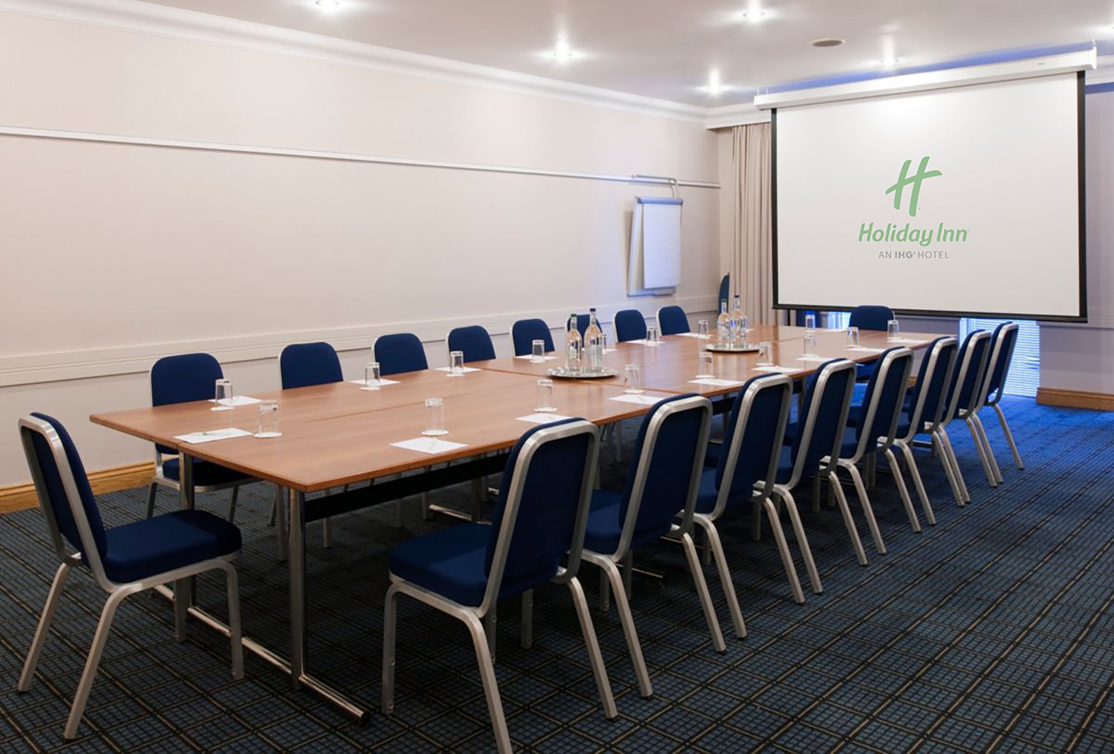 A conference room with a long table, blue chairs, and a projector screen displaying the Holiday Inn logo.