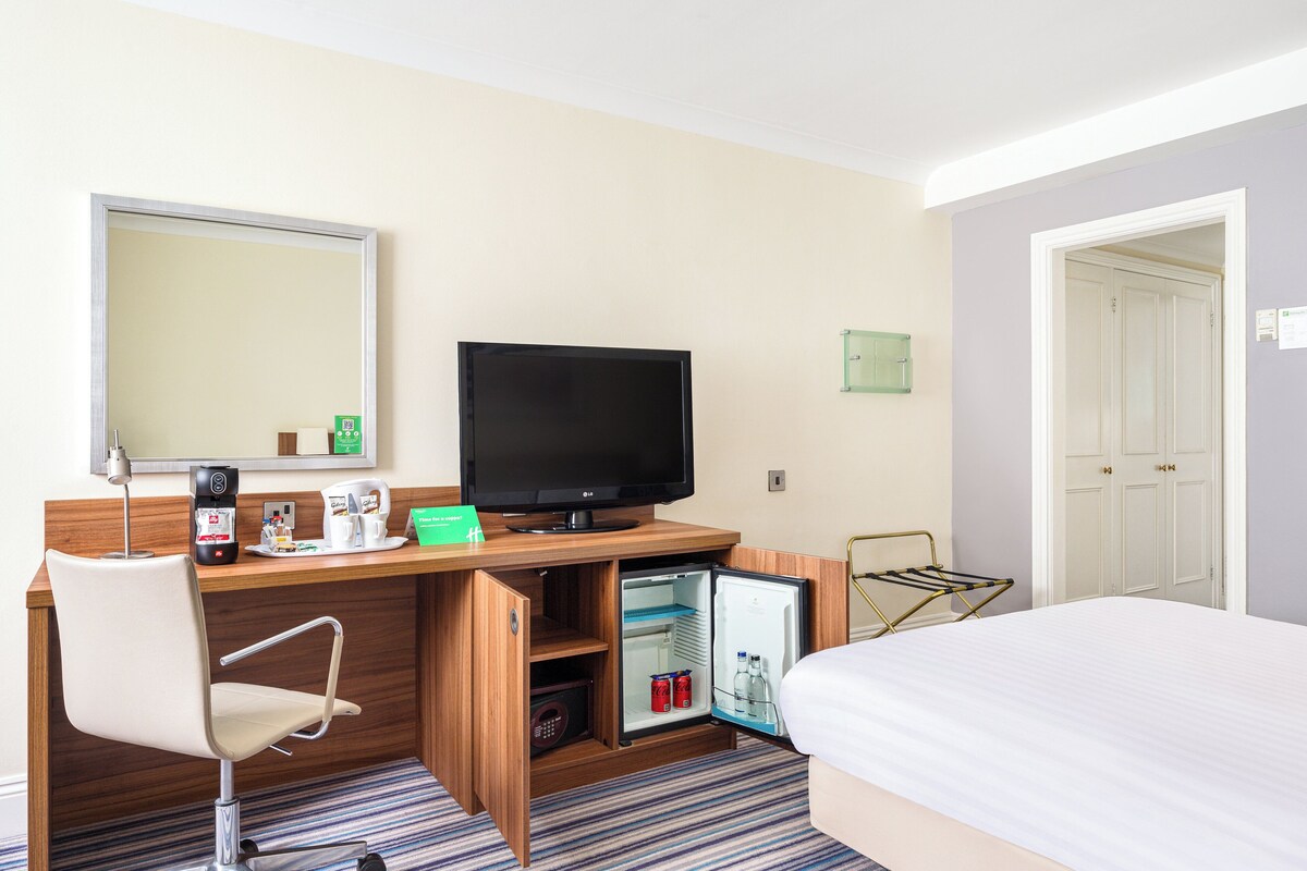 A hotel room with a bed, a desk with a television, a mini-fridge, and a chair.