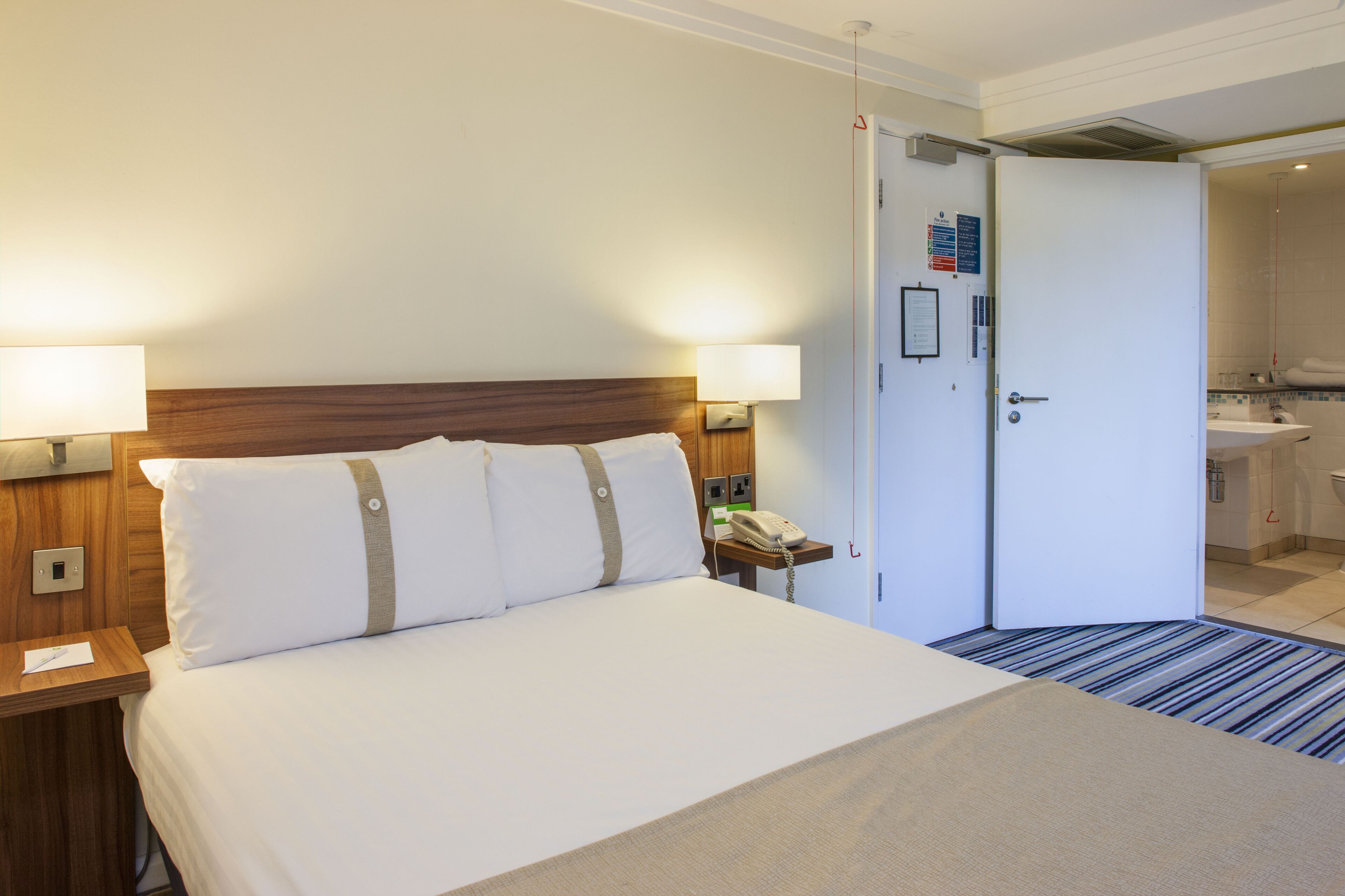 Una camera d'albergo con un letto, abat-jour, un telefono e una porta che dà su un bagno.