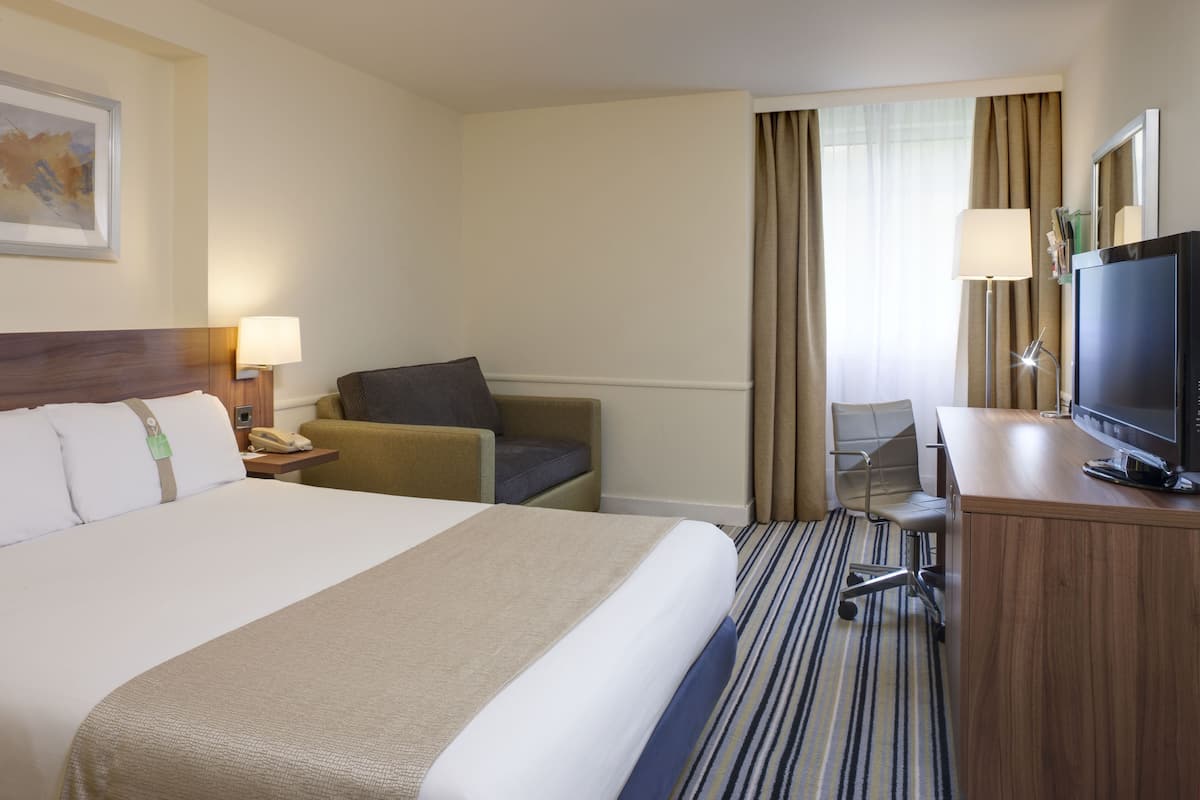 A hotel room with a large bed, a desk with a television, a chair, and a lamp.