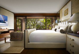 A modern bedroom with a large bed, a desk, and a view of lush greenery through large windows.