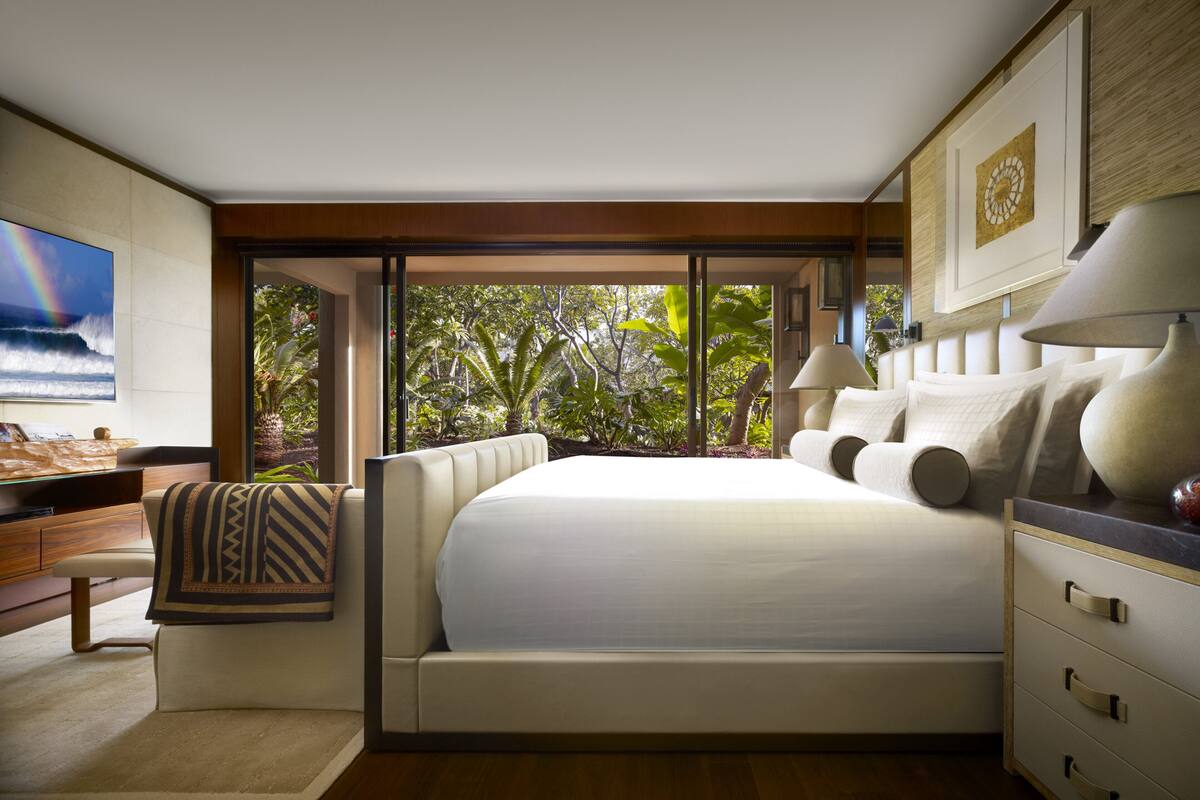 Ein modernes Schlafzimmer mit einem großen Bett, einem Schreibtisch und Blick auf üppiges Grün durch große Fenster.