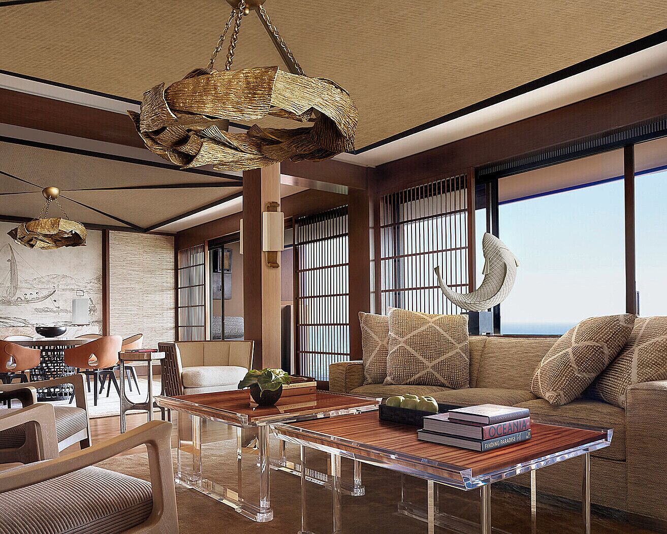A spacious living area with a large sofa, a coffee table, and a dining table. There are large windows providing a view of the ocean.